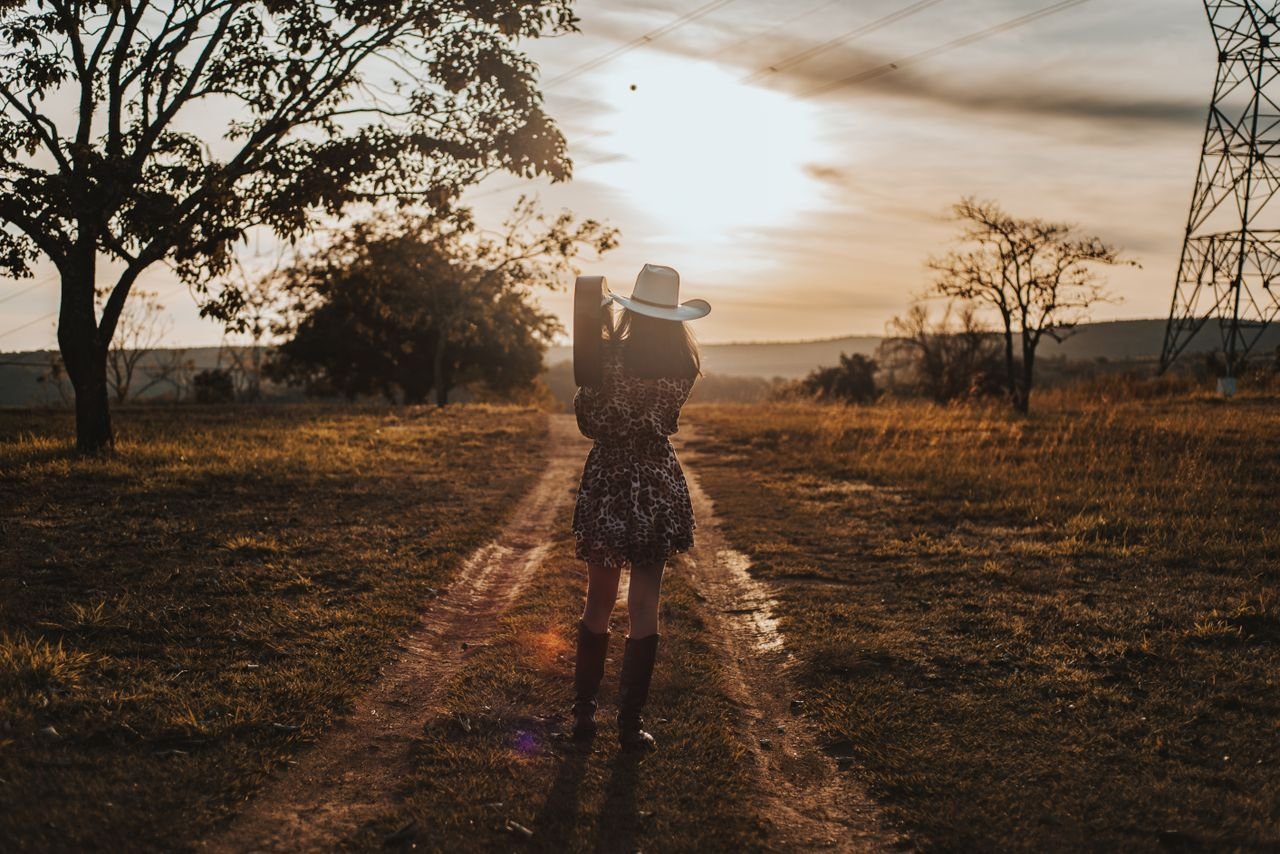 A woman stands in a field at sunset, wearing a leopard print dress and white hat.